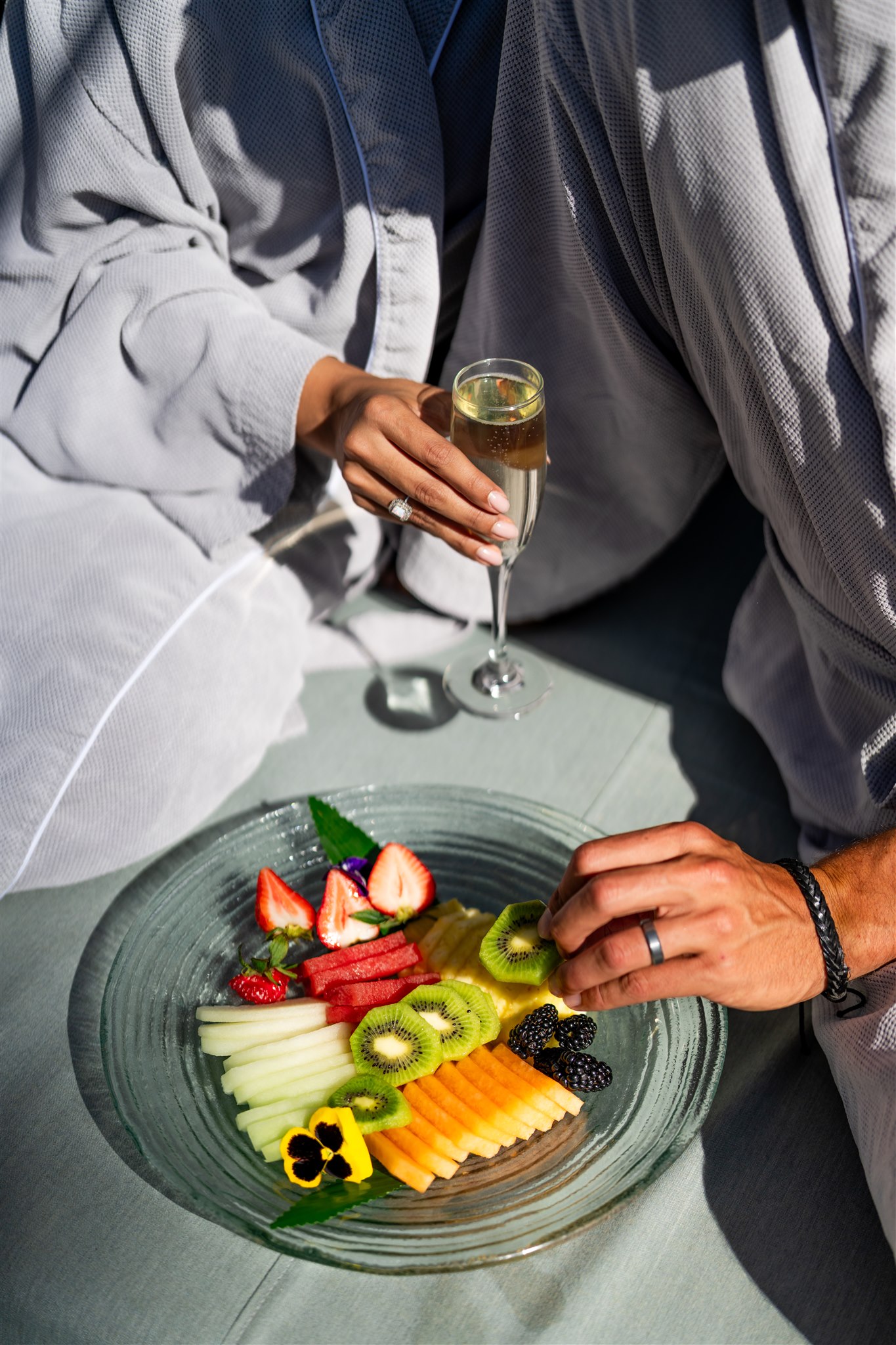 a person in a robe holding a glass of champagne next to a plate of fruit