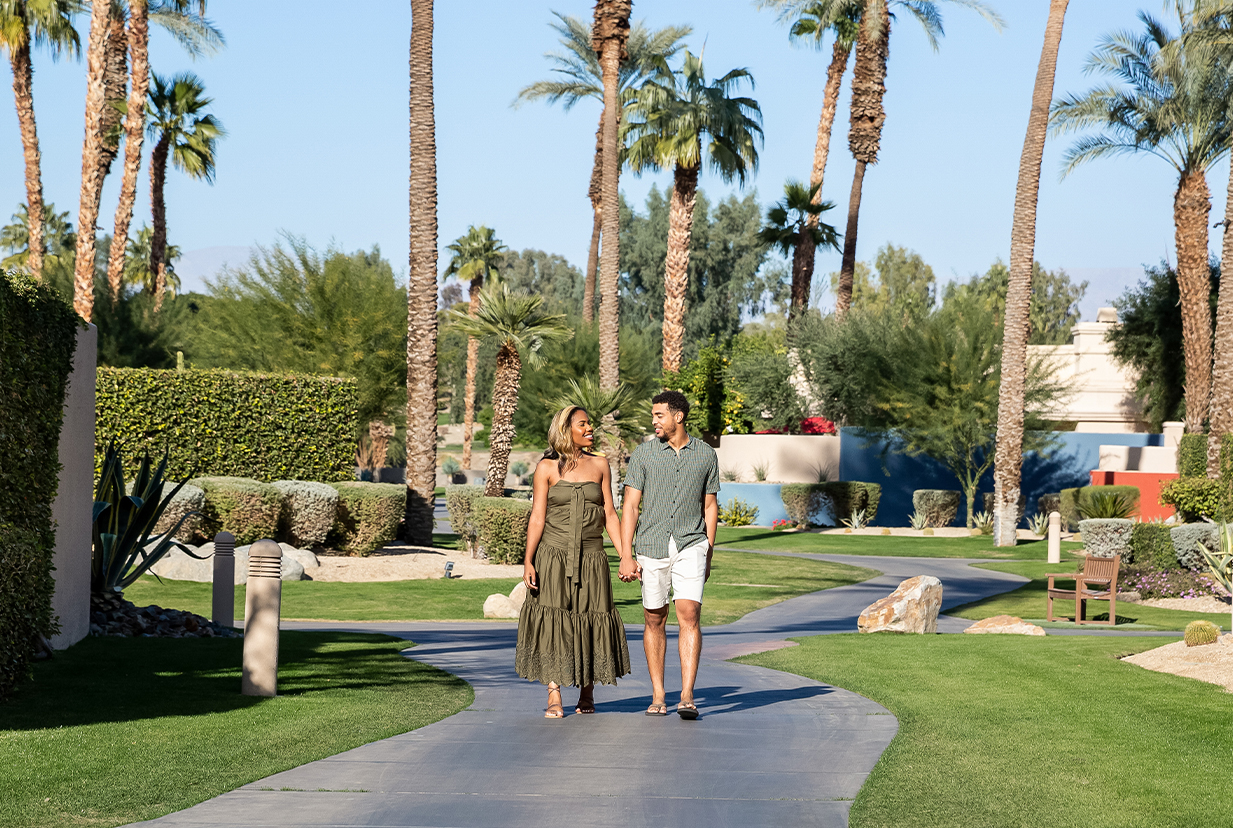a man and woman holding hands walking on a path with palm trees
