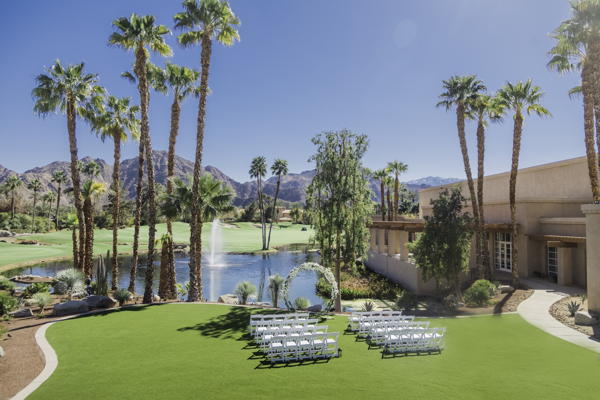 a lawn with white chairs and a fountain in front of a building