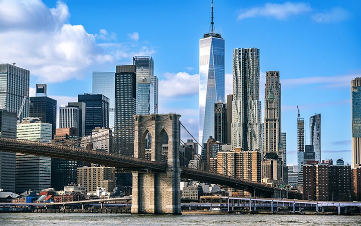 A city skyline with a bridge in the foreground and skyscrapers in the background under a blue sky with clouds.