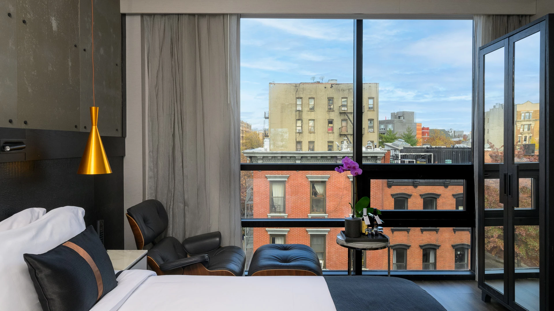 A hotel room with a bed, chair, and table in front of a large window showing a city view.