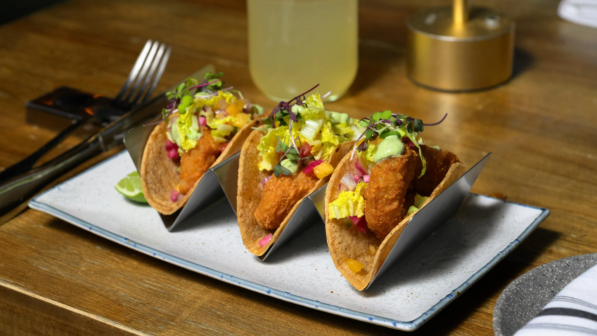 A plate of tacos on a wooden table with utensils and a drink