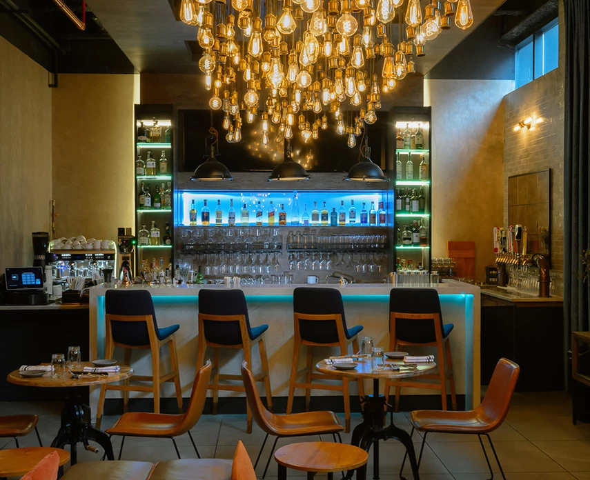 A dimly lit bar with a large chandelier, barstools, and tables.
