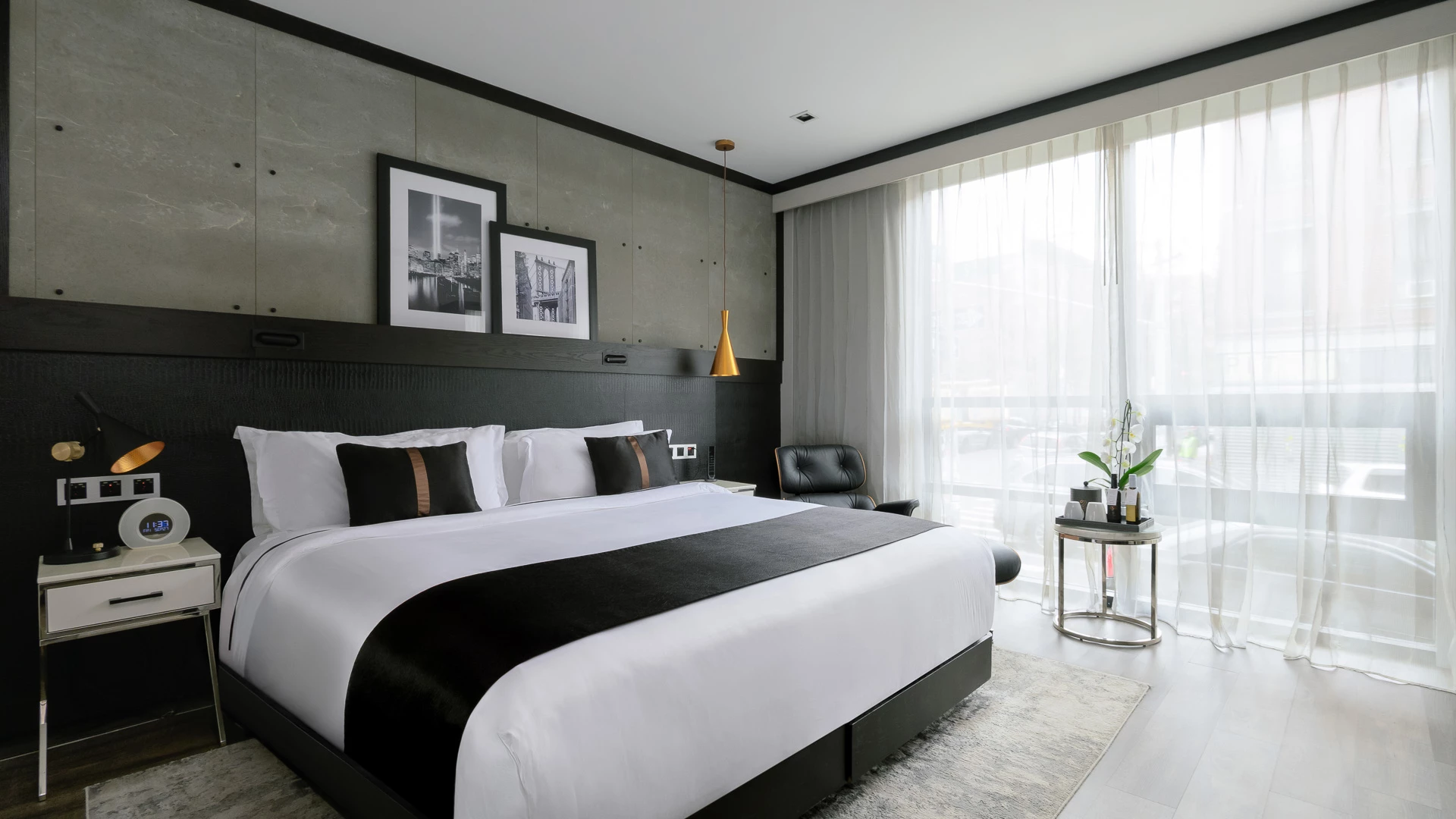 A modern hotel room with a large bed, nightstand, chair, and window with sheer curtains.
