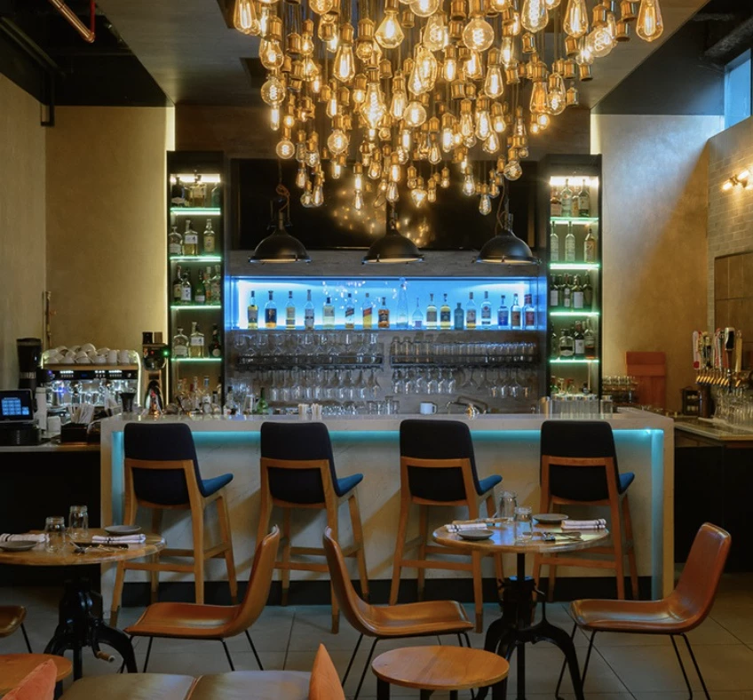 A dimly lit bar with a large chandelier, shelves of bottles, and several tables with chairs.