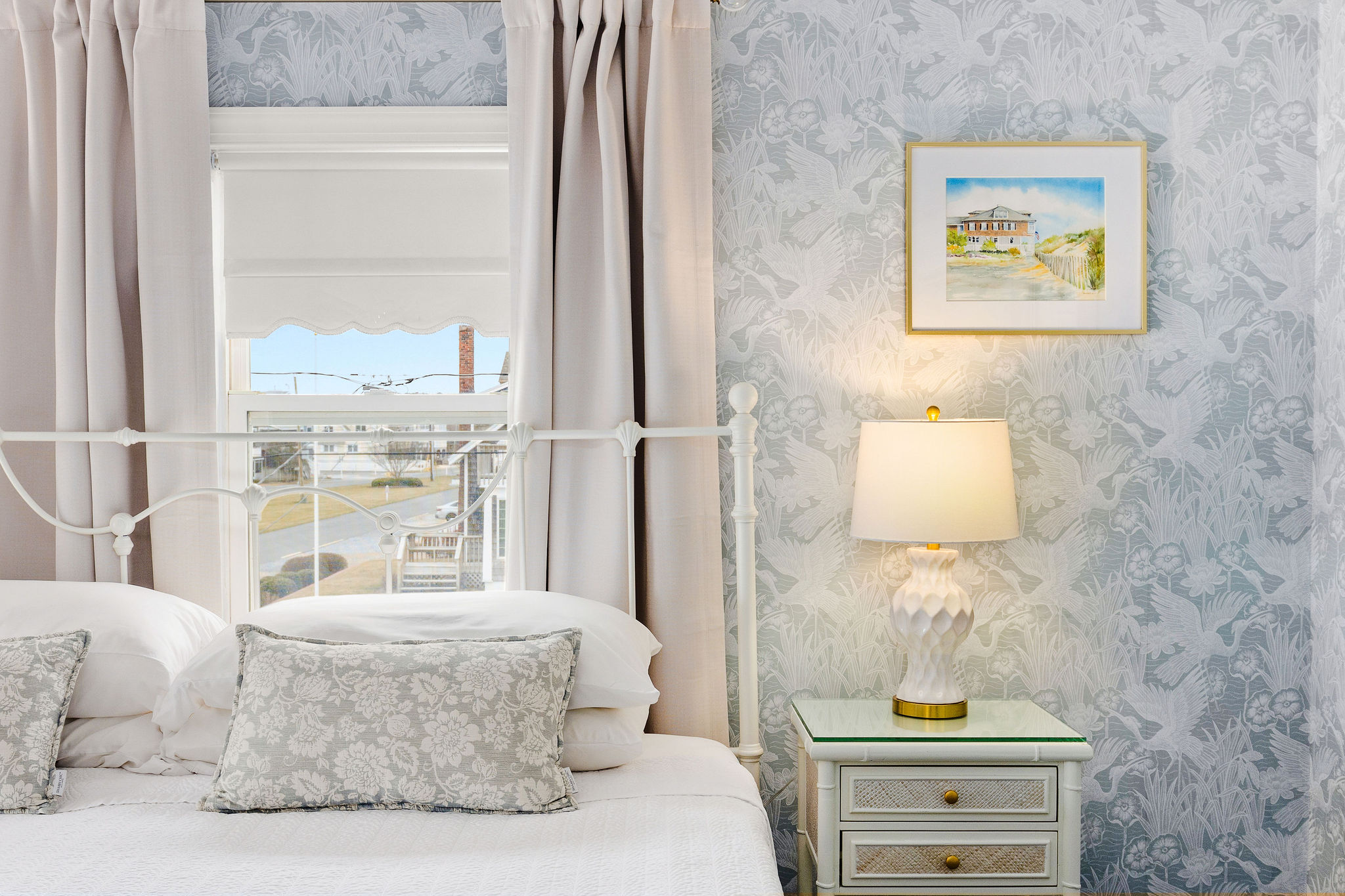 A neatly made bed with white linens and gray pillows in a room with a window, nightstand, and artwork.