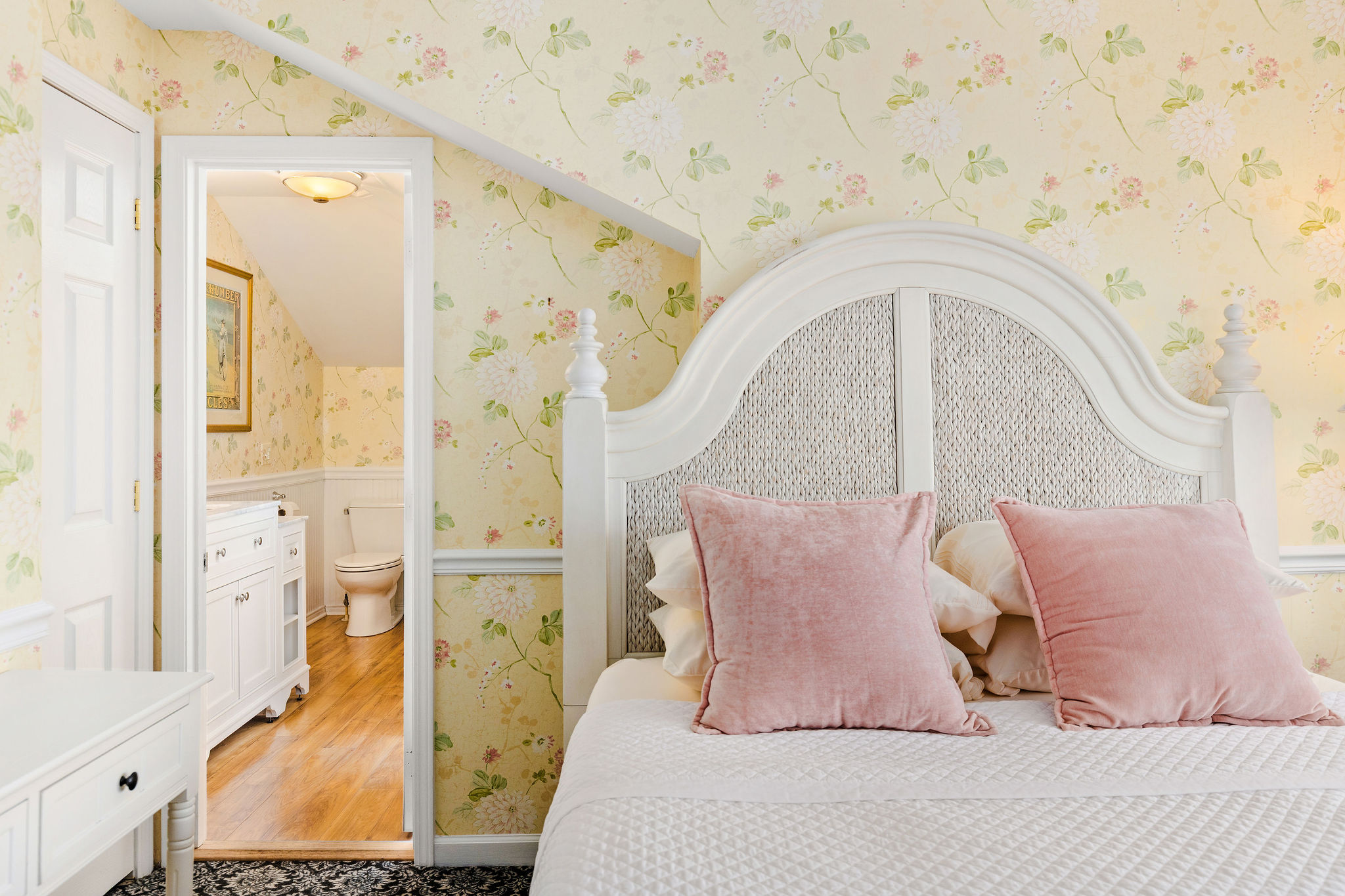 A white bed with pink pillows in a room with floral wallpaper and a doorway to a bathroom.