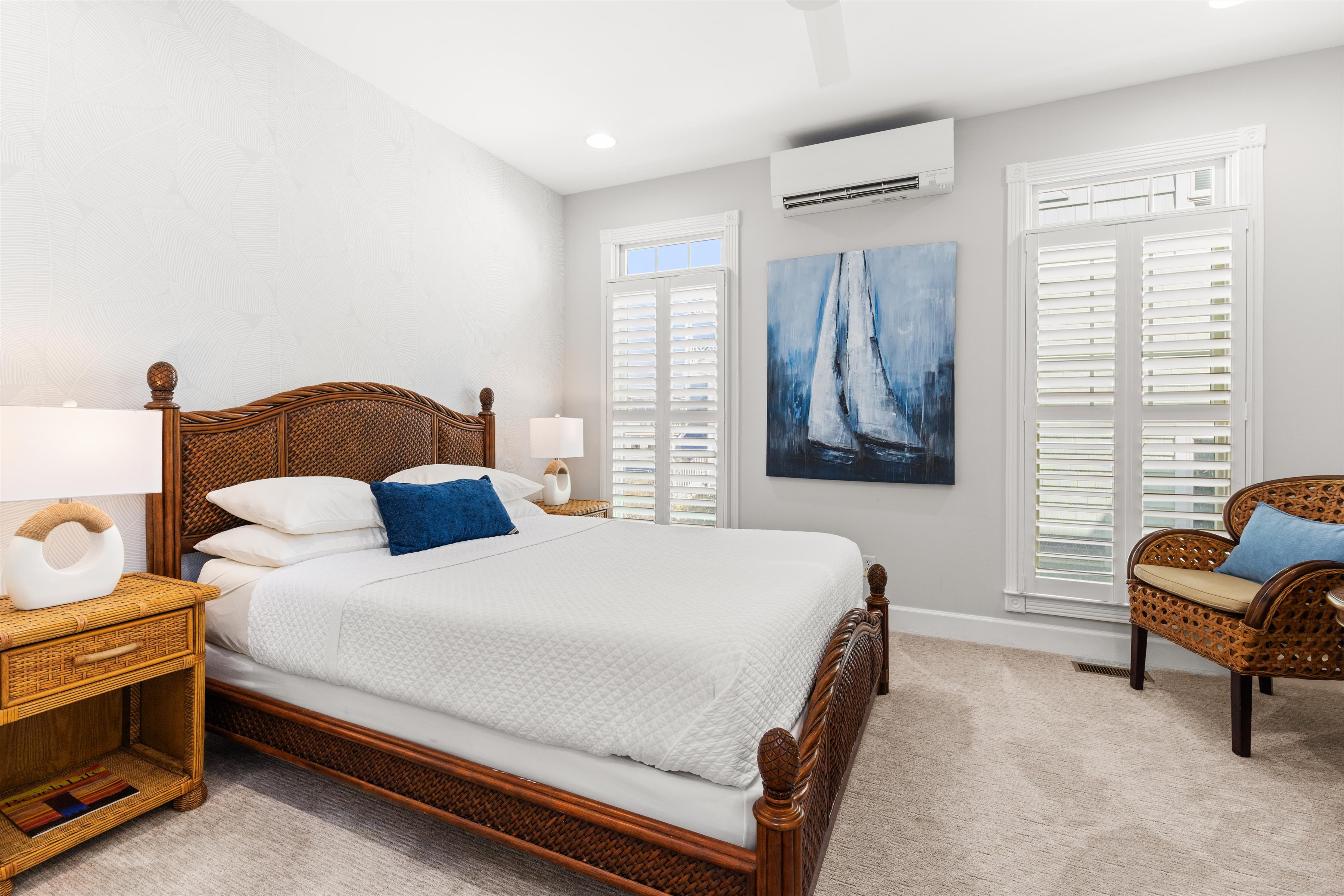A wicker-framed bed with white bedding and blue accents sits in a bright, airy room with white walls and beige carpet.