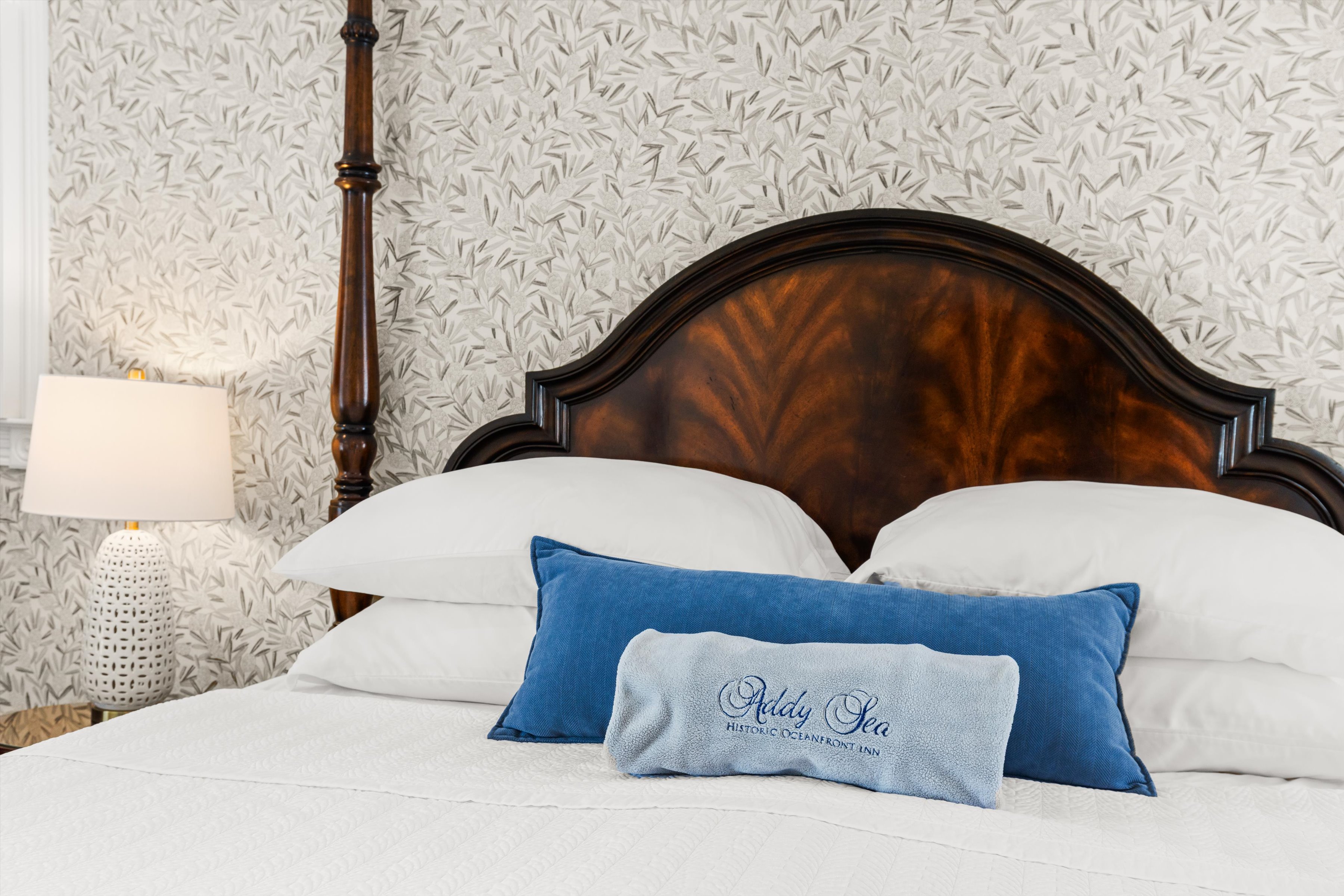 A neatly made bed with a wooden headboard, white bedding, and blue accent pillows, accompanied by a lamp on a nightstand, in a room with patterned wallpaper.
