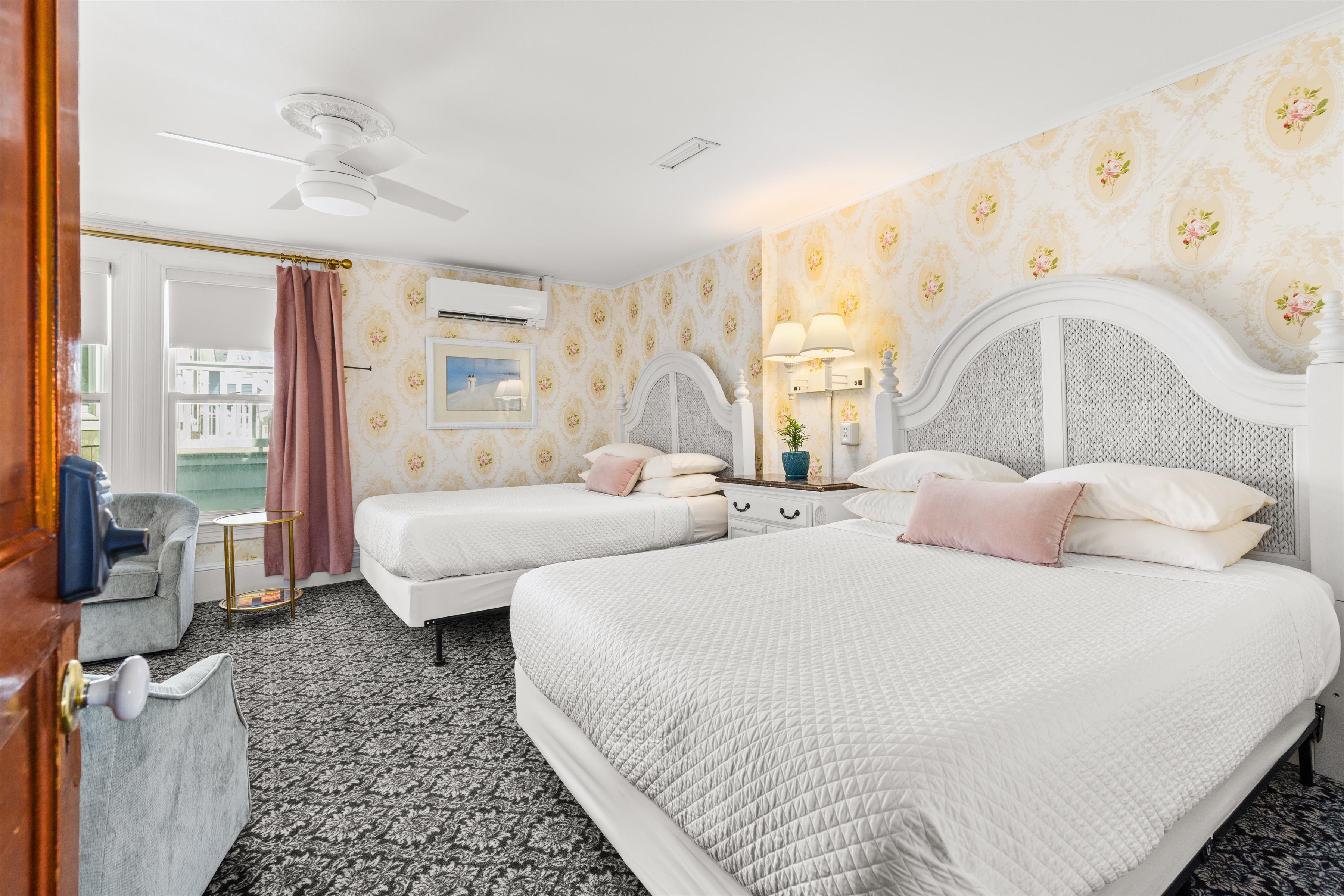 A wide view of a hotel room with two beds, floral wallpaper, and a window with a view.