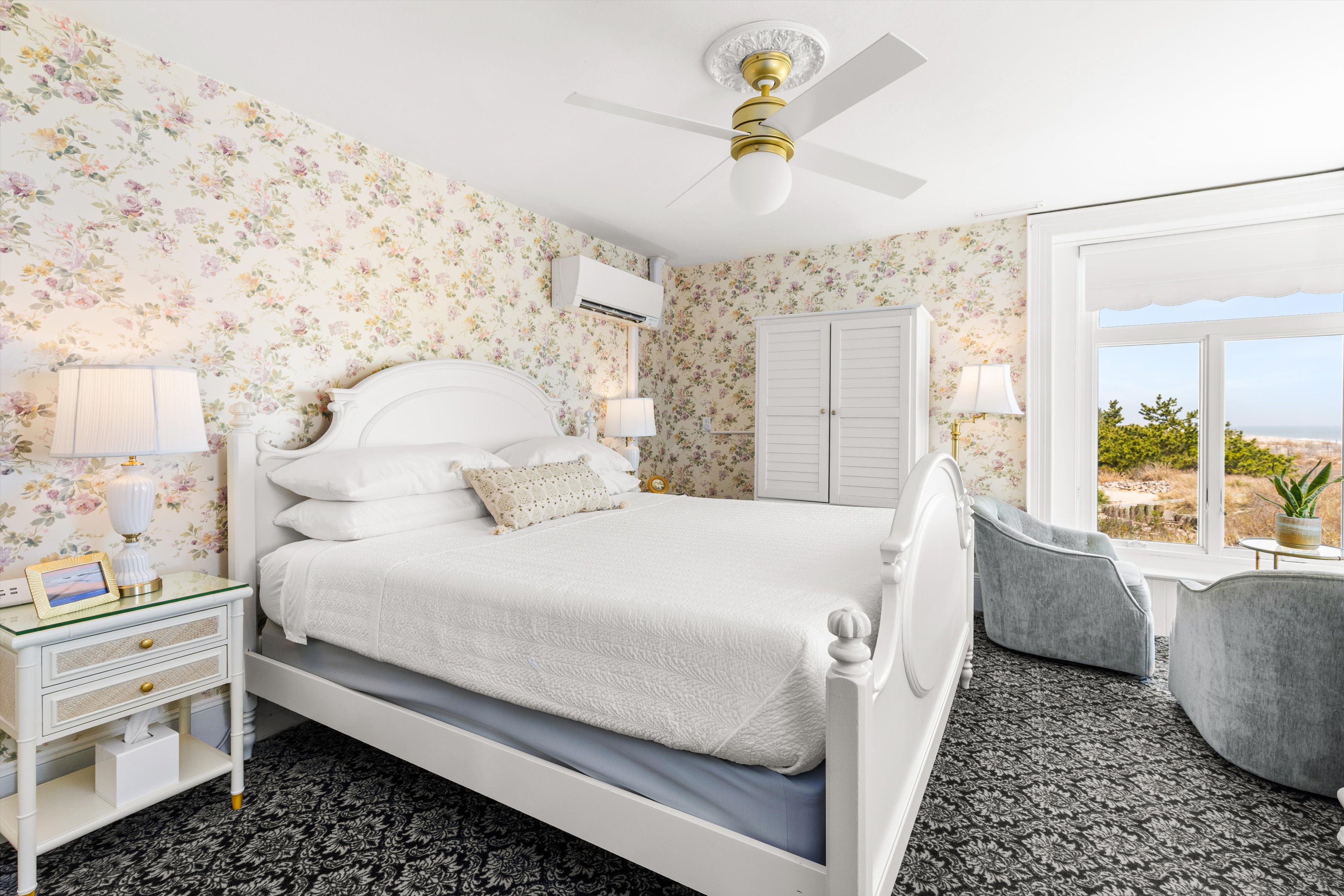 A wide view of a guest room with a white bed, floral wallpaper, and a large window.