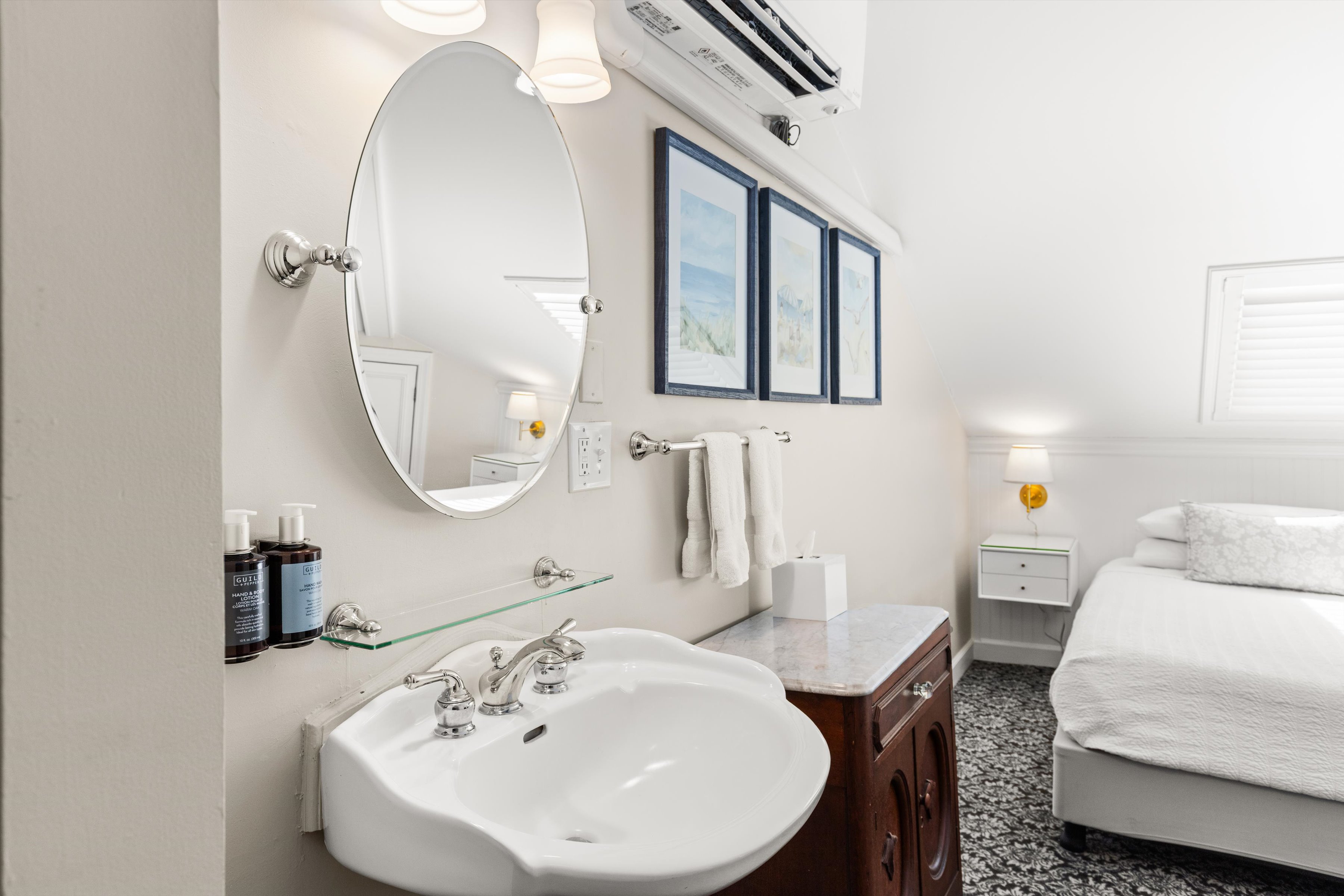 A clean and well-lit bathroom with a white sink, round mirror, and a bed visible in the background.