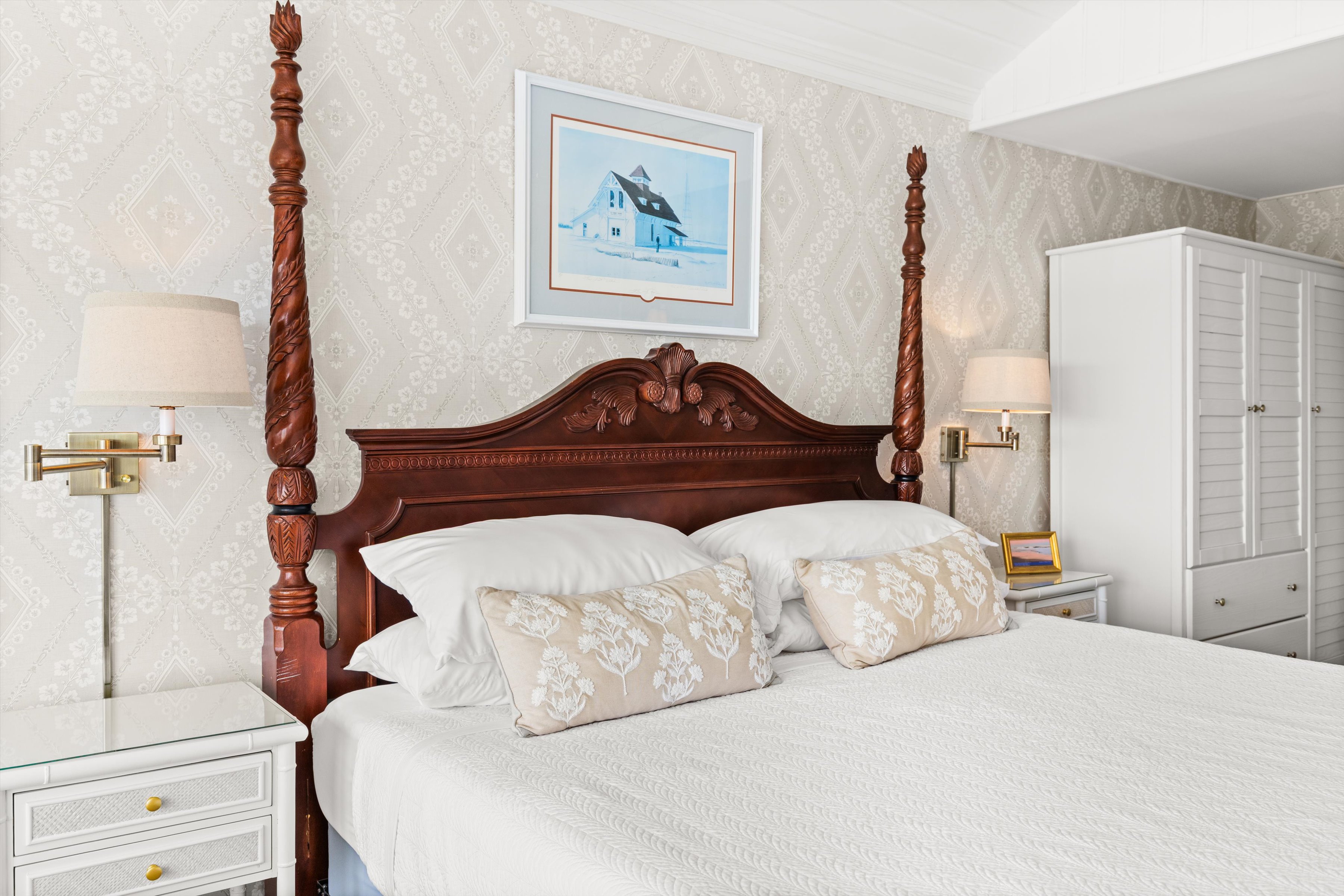 A neatly made bed with a dark wooden four-poster frame, white bedding, and beige pillows in a room with a wardrobe and lamps.