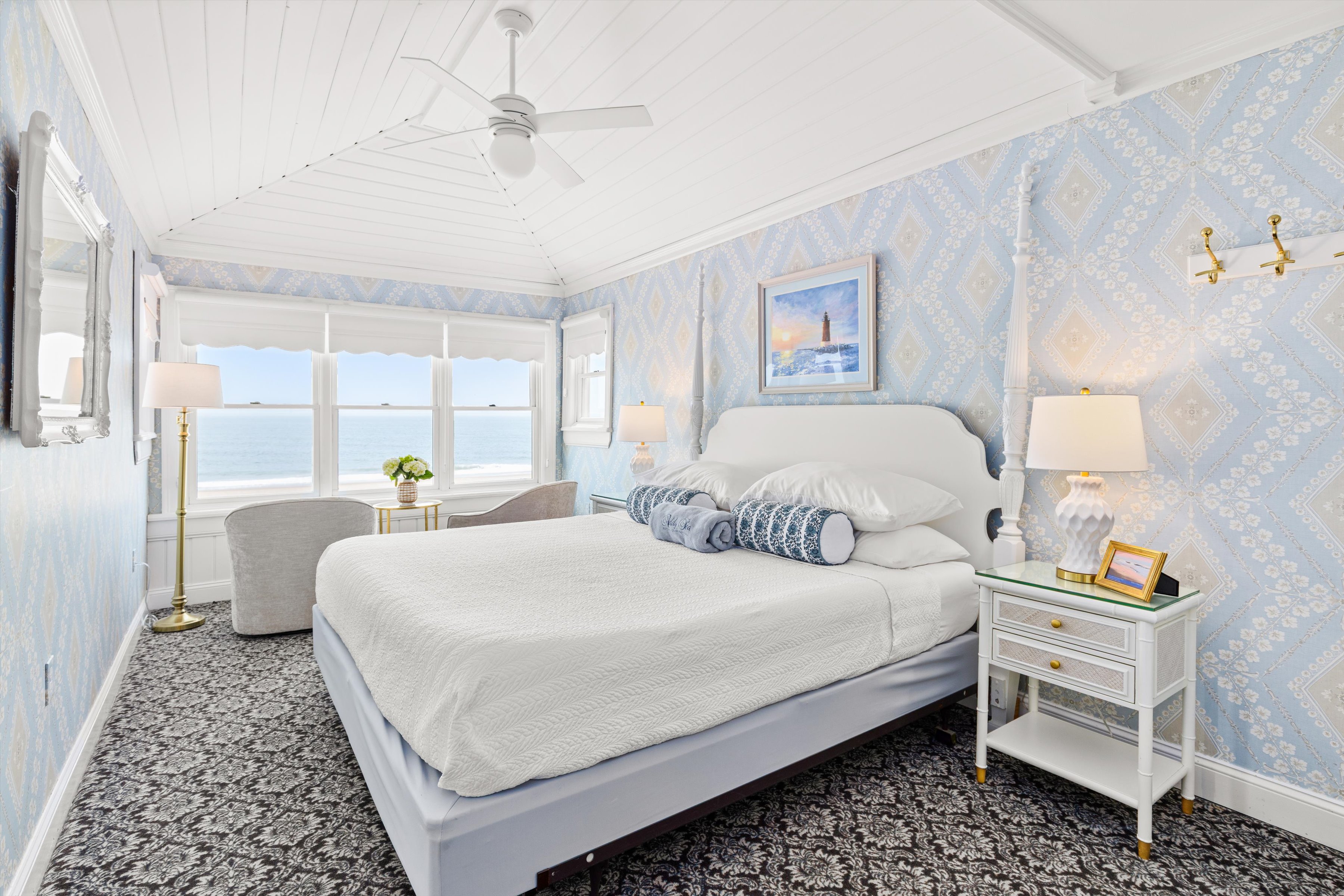 A wide-angle view of a guest room with a white bed, blue wallpaper, and an ocean view.