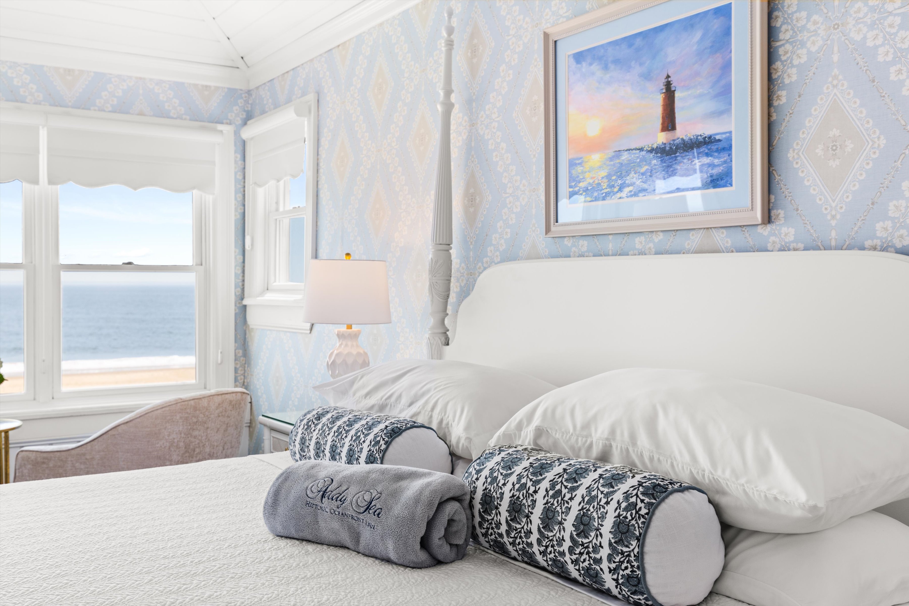 A neatly made bed in a room with blue wallpaper, a window overlooking the sea, and a piece of artwork on the wall.