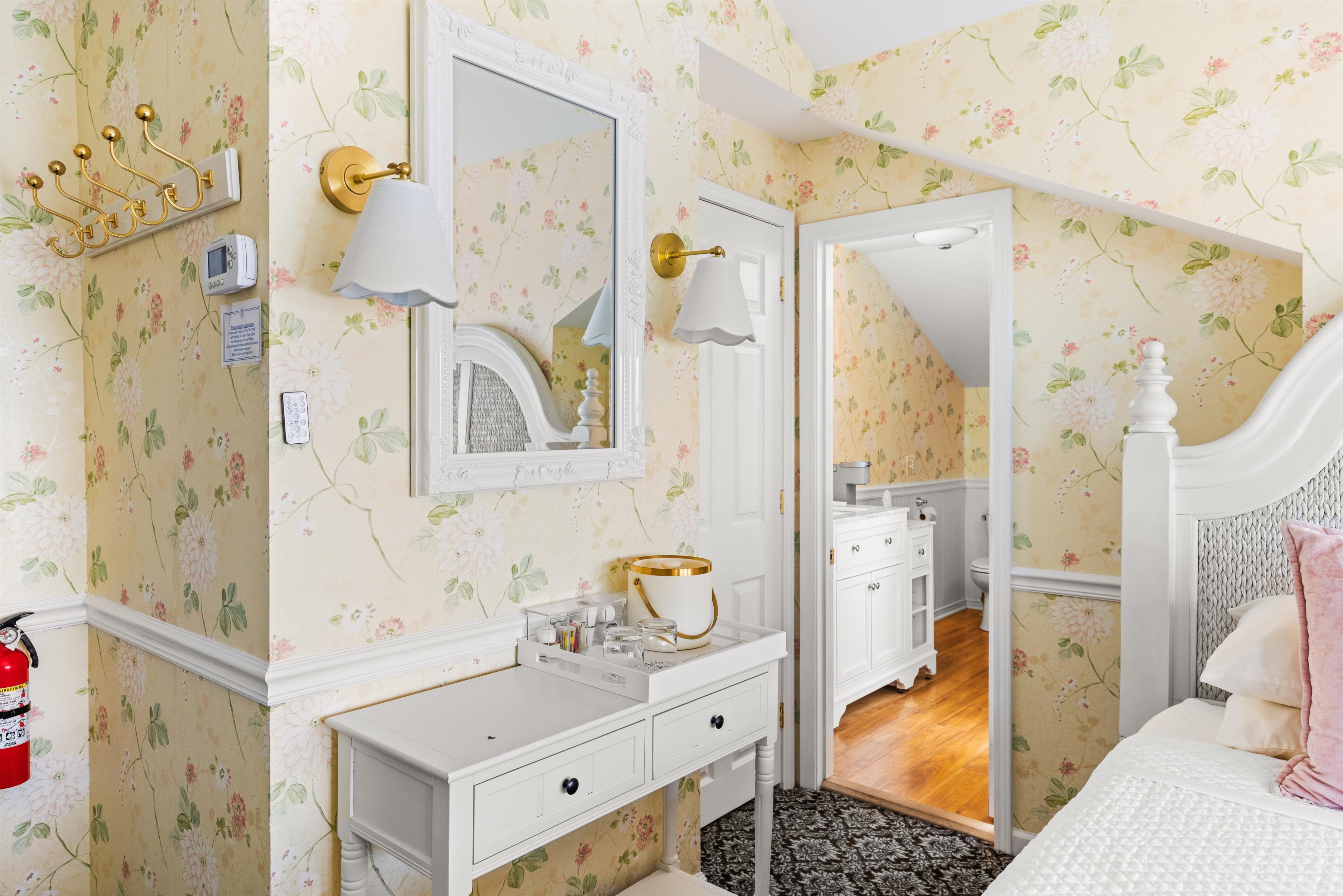 A wide-angle view of a hotel room with floral wallpaper, a white bed, and a vanity area.
