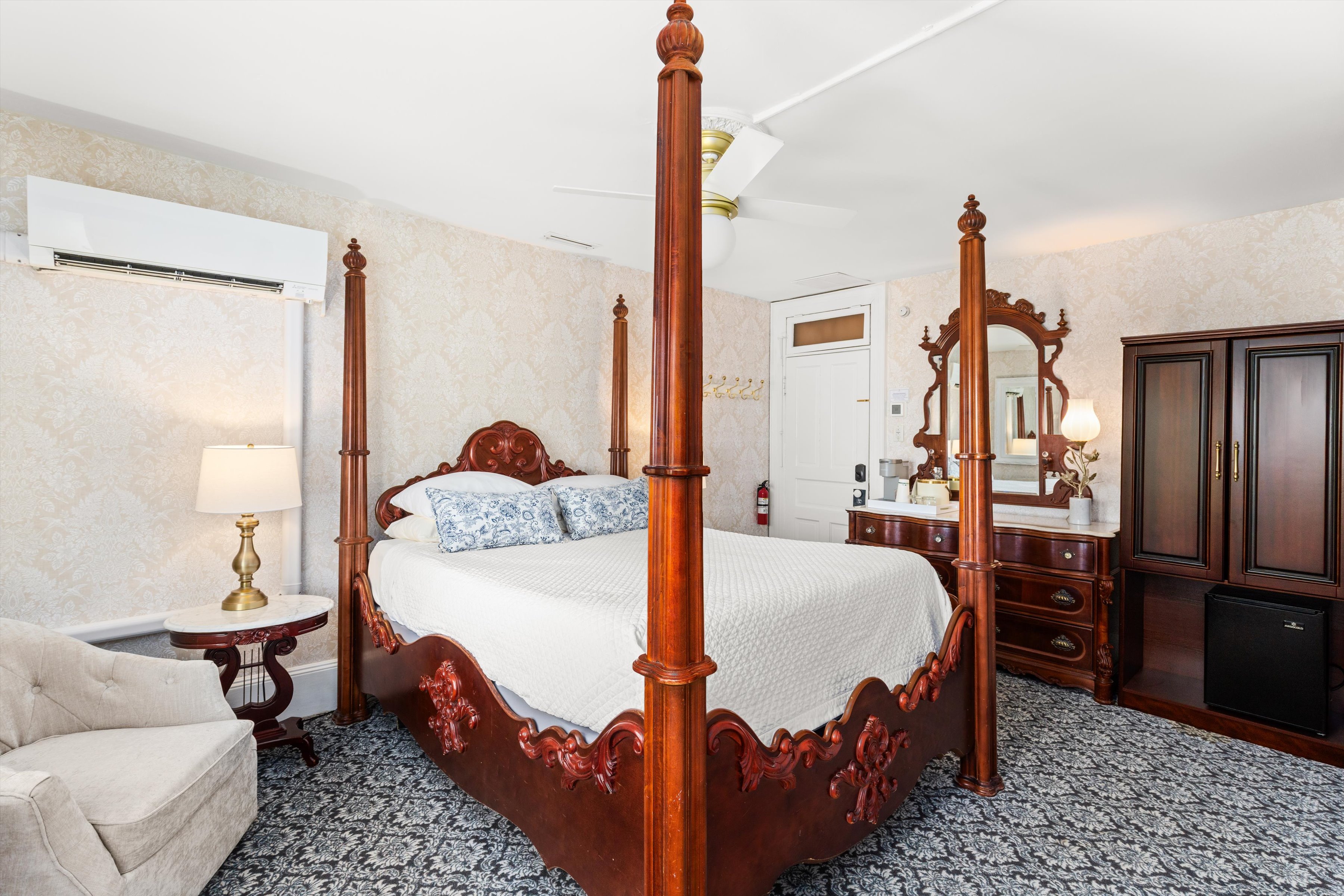 A wide-angle view of a guest room with a four-poster bed, dresser, armchair, and ceiling fan.