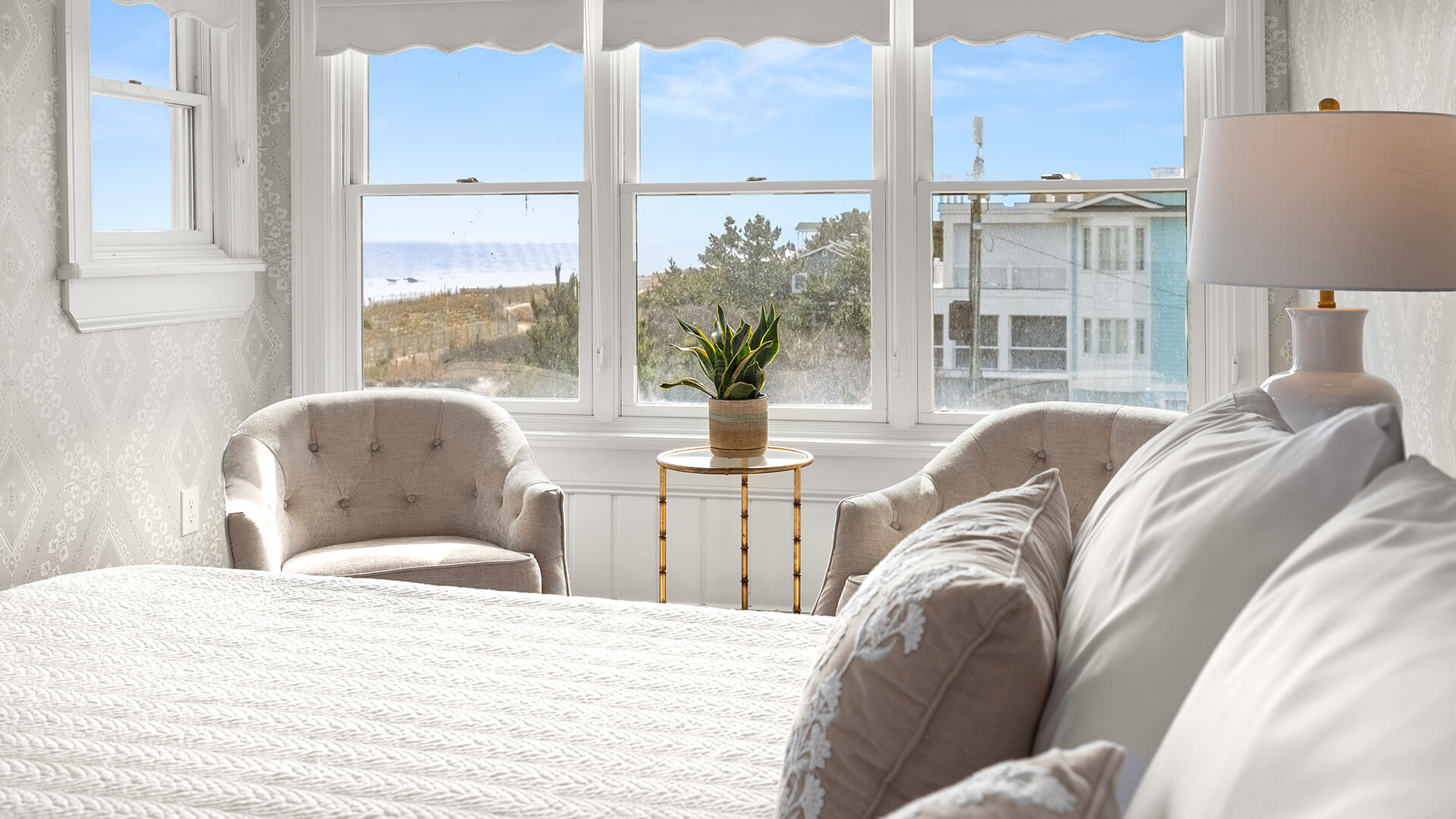 A guest room with a large window showing an ocean view, a bed, two chairs, and a table with a lamp.