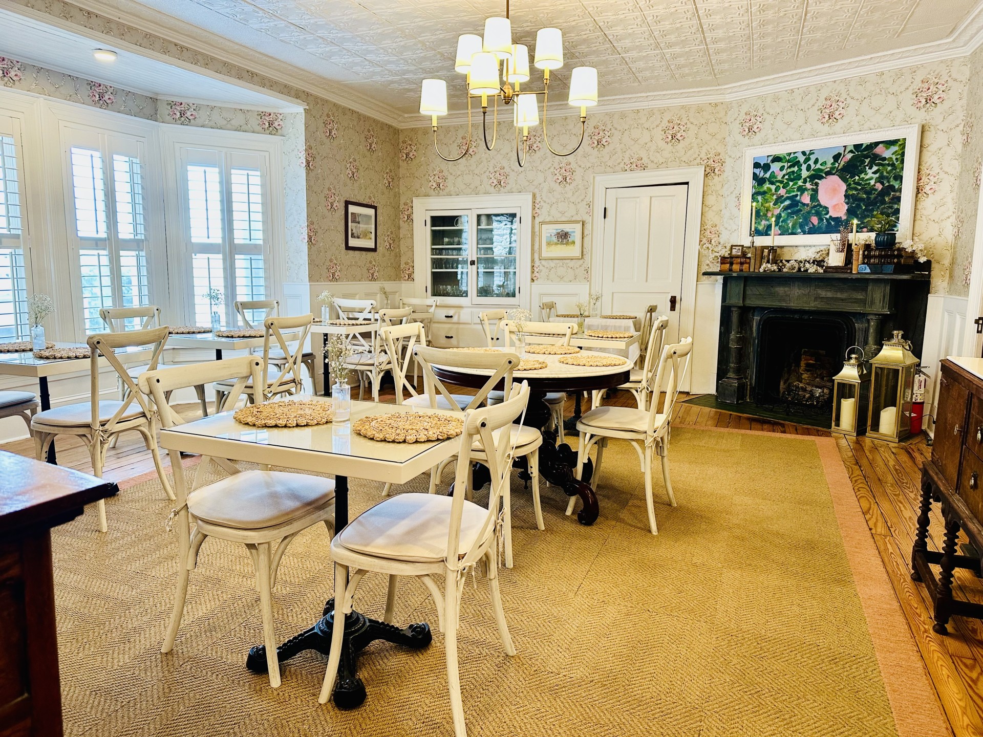 A brightly lit dining room with white chairs, floral wallpaper, and a fireplace.