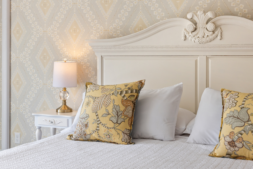 A bed with white sheets and yellow pillows, a lamp on a nightstand, and wallpaper on the wall.