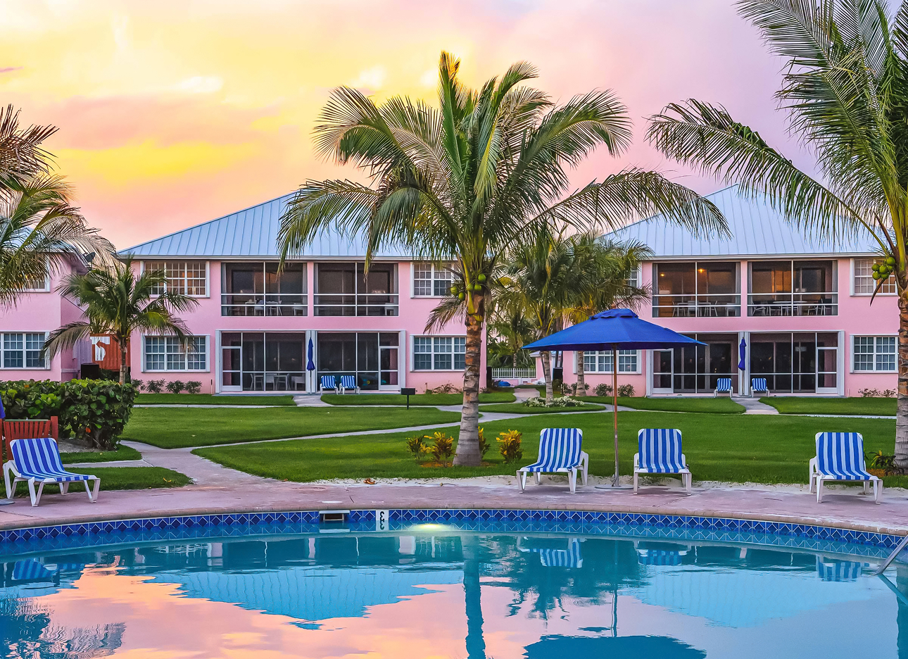 a pool with lawn chairs and palm trees in front of a pink building