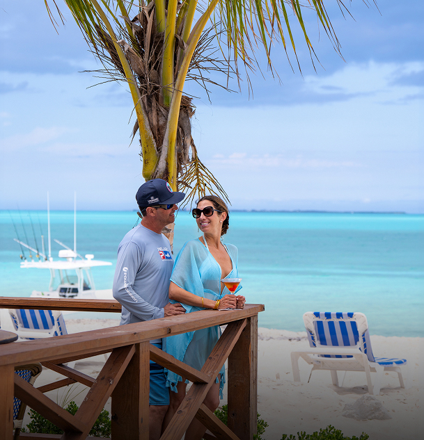 a man and woman standing on a deck with a palm tree and chairs on the beach
