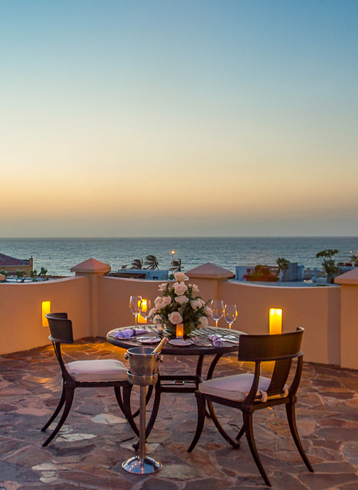 a table set up with wine glasses and flowers on a patio overlooking the ocean