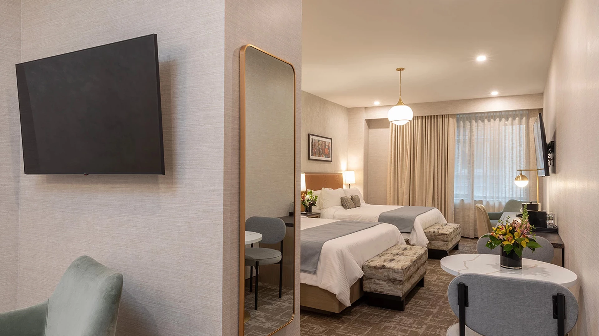 A hotel room with two beds, a desk, a table with chairs, and a TV on the wall.