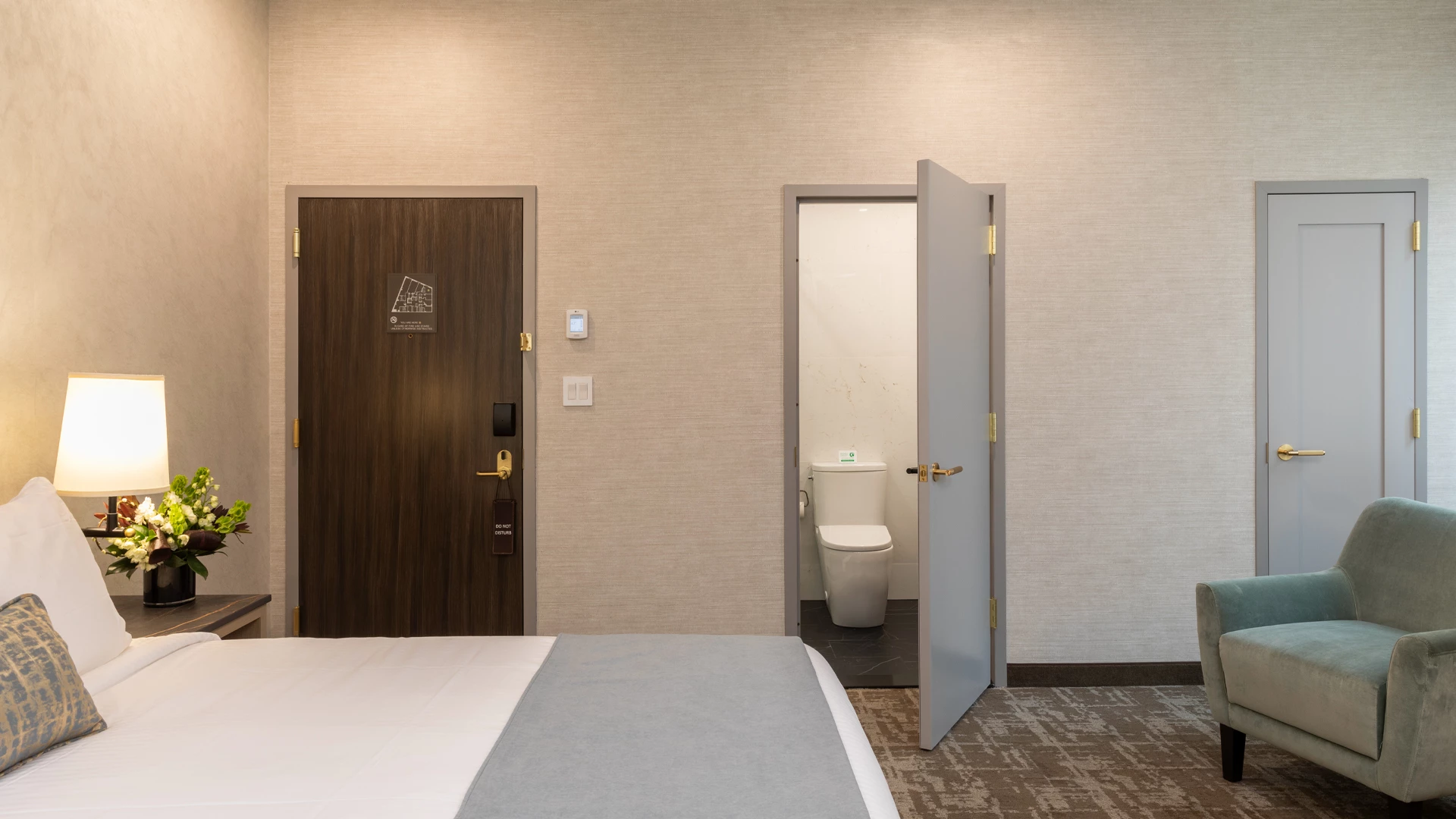 A hotel room with a bed, nightstand, lamp, flowers, chair, and bathroom