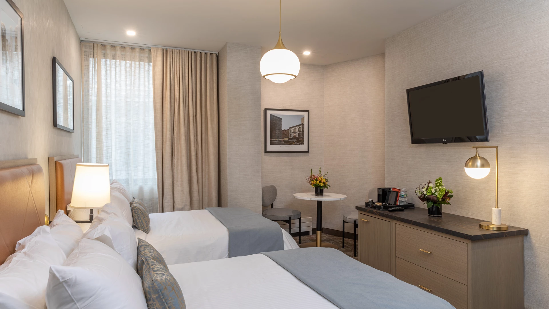 A hotel room with two beds, a TV, a table with flowers, and a lamp.