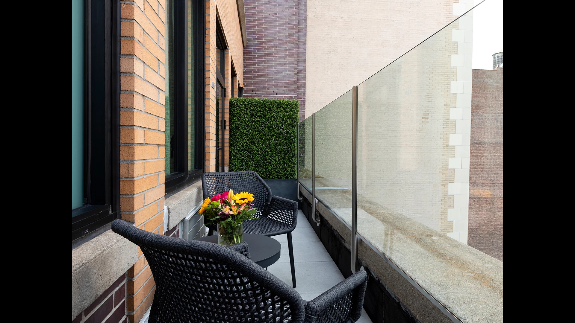 A small balcony with a glass railing, two wicker chairs, and a vase of flowers.