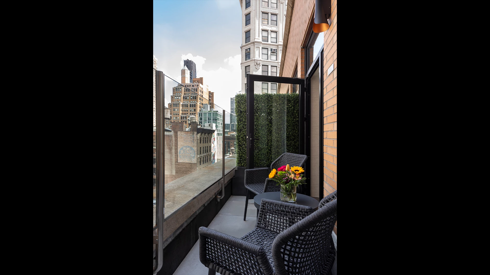 A balcony with a glass railing, wicker chairs, and a vase of flowers, overlooking a cityscape.