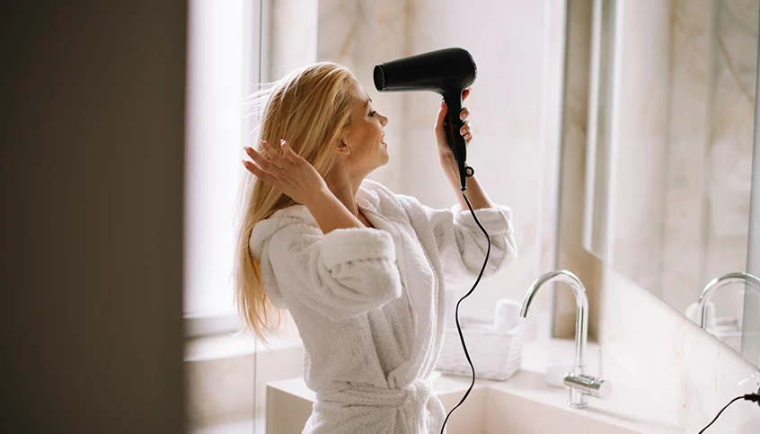 A woman in a white robe is drying her hair with a black hair dryer in front of a marble sink and mirror.