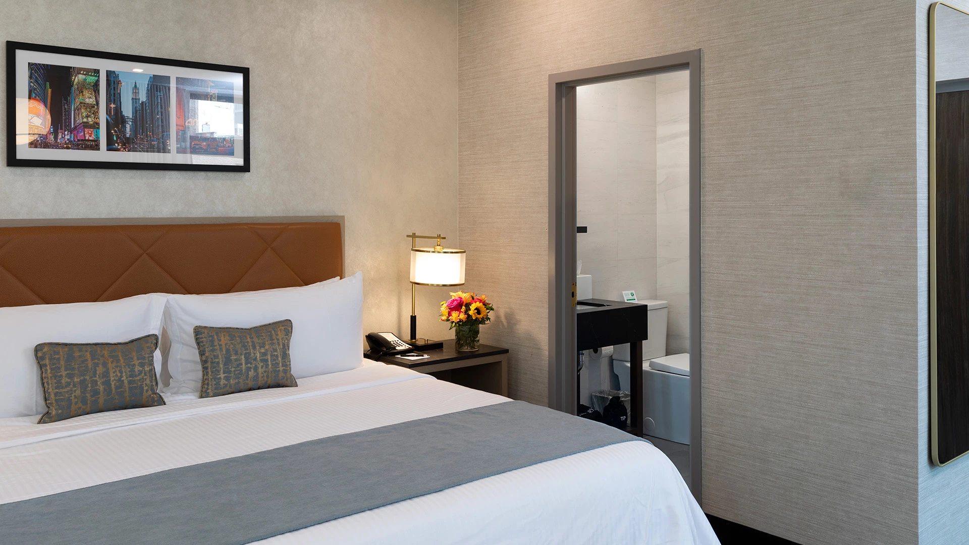 A hotel room with a bed, nightstand, lamp, and artwork