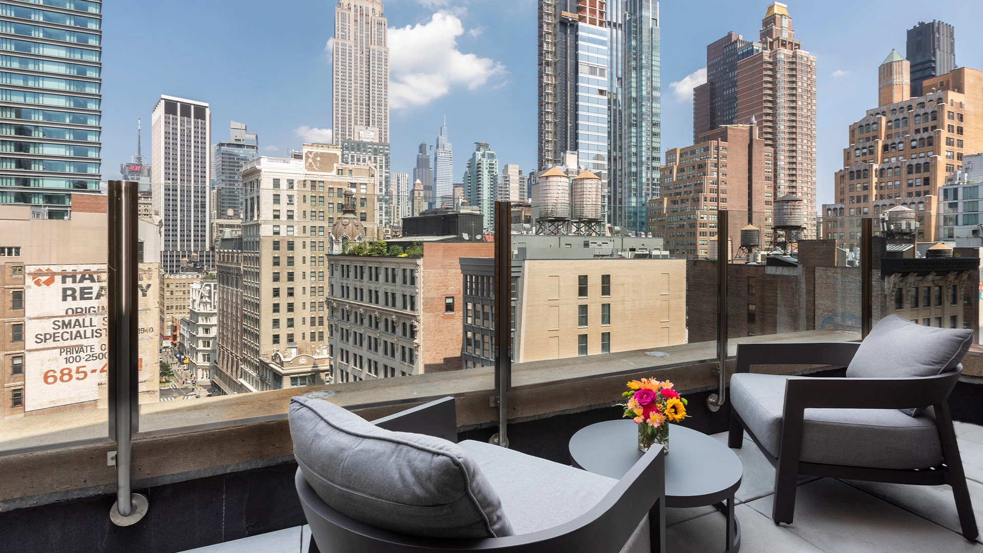 A balcony with a city view, featuring two chairs, a table, and flowers.