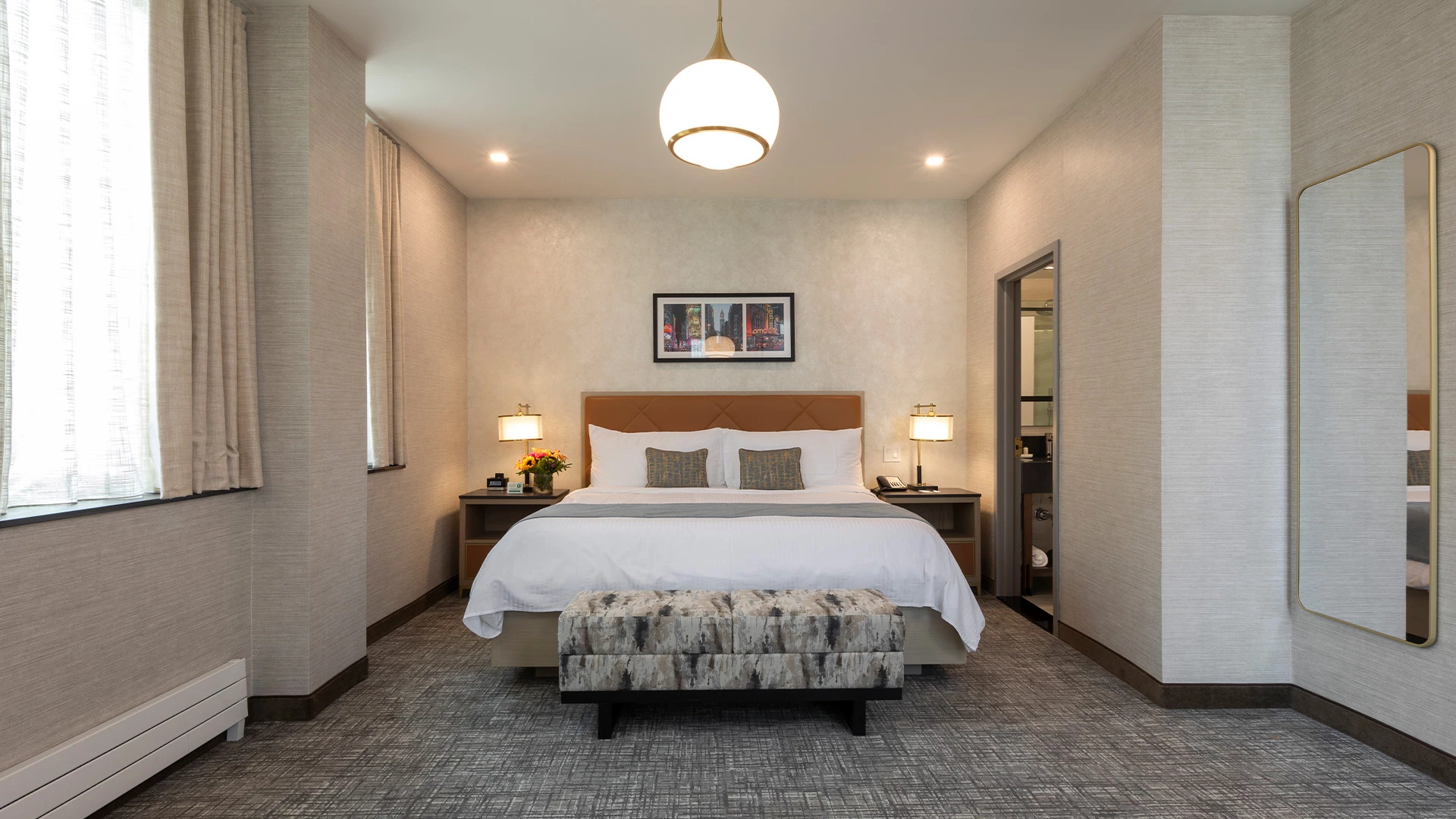 A hotel room with a large bed, a bench at the foot of the bed, two nightstands with lamps, a window with curtains, and a mirror on the wall.