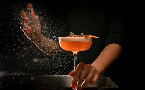 A hand holding a cocktail glass with a pink drink and a sprig of herbs, with another hand sprinkling a garnish.