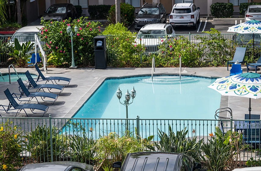 A fenced pool area with lounge chairs and umbrellas, surrounded by plants and a parking lot.