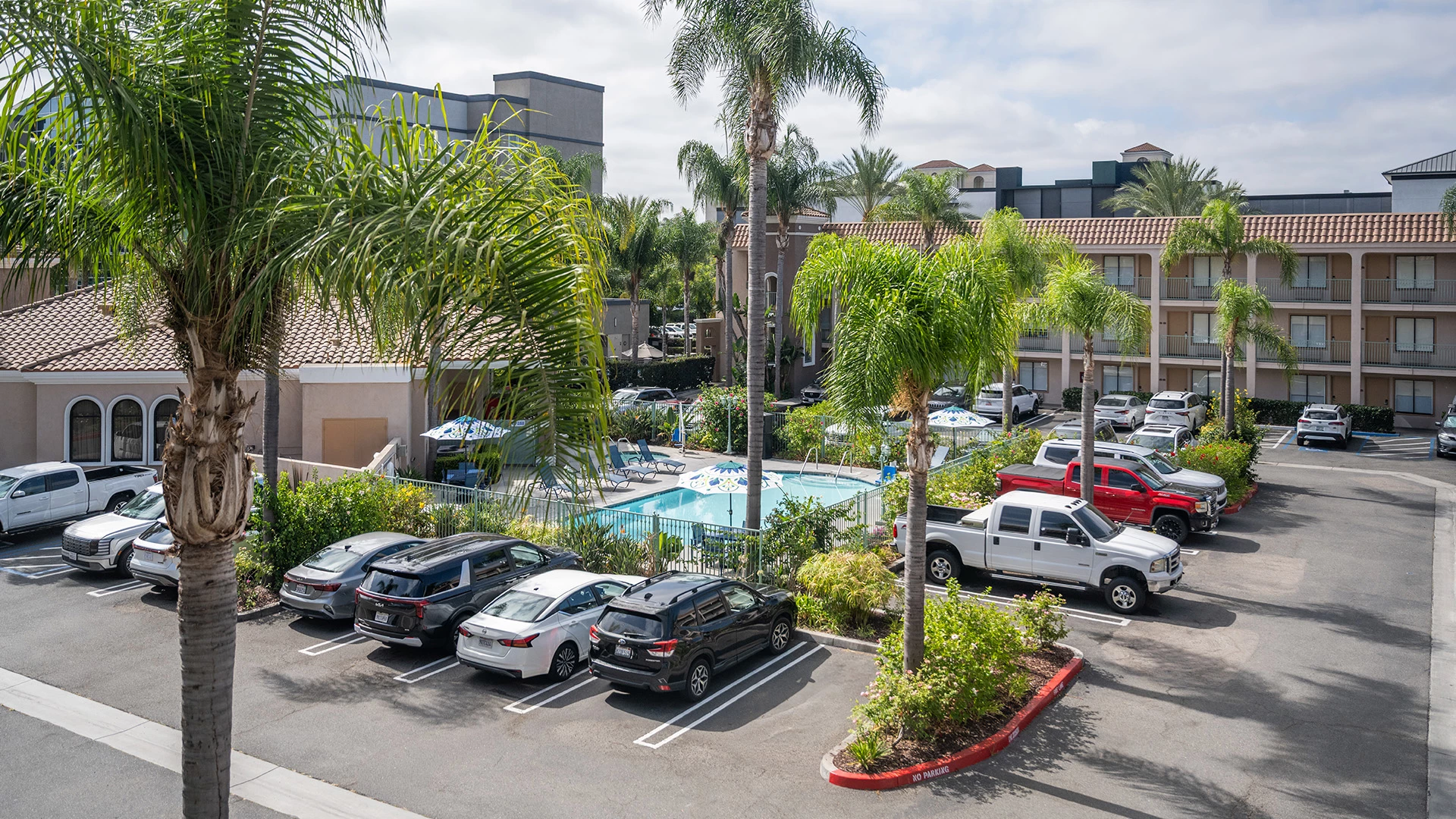 A parking lot with cars and palm trees in front of a hotel building with a pool.