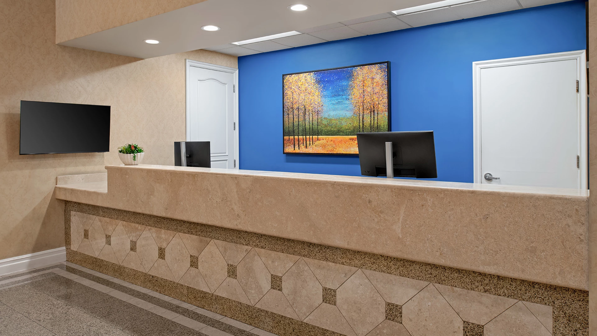 A hotel lobby with a reception desk, two computer monitors, a TV, and a painting on the wall.