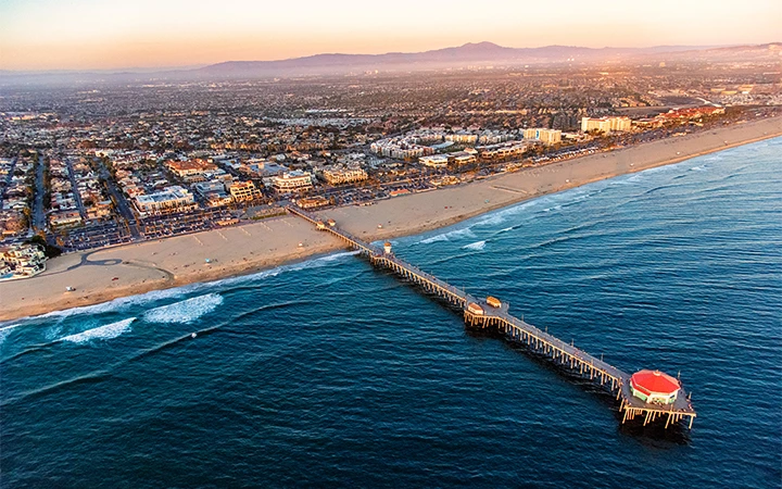 Aerial view of a beach with a long pier extending into the ocean, with buildings and mountains in the background.