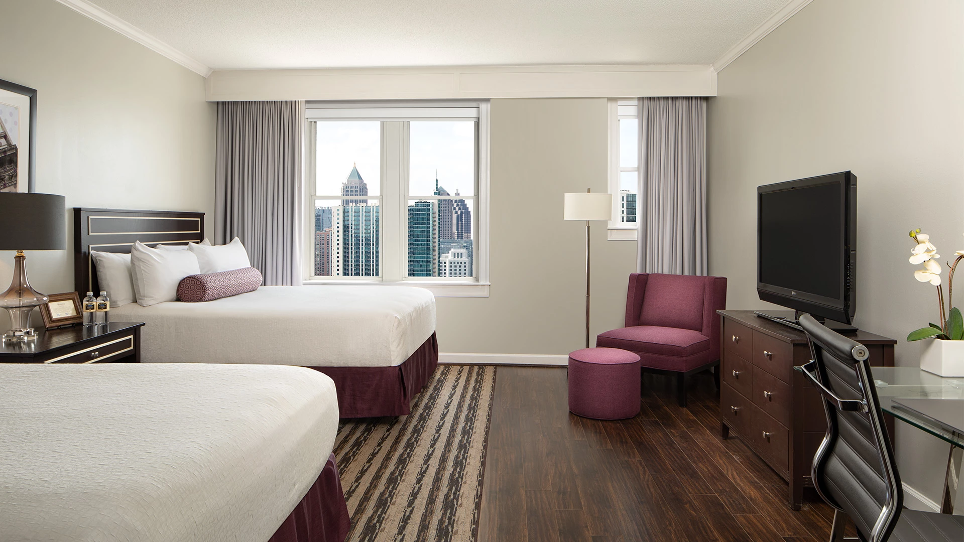 A hotel room with two beds, a desk, TV, and a window with a city view.