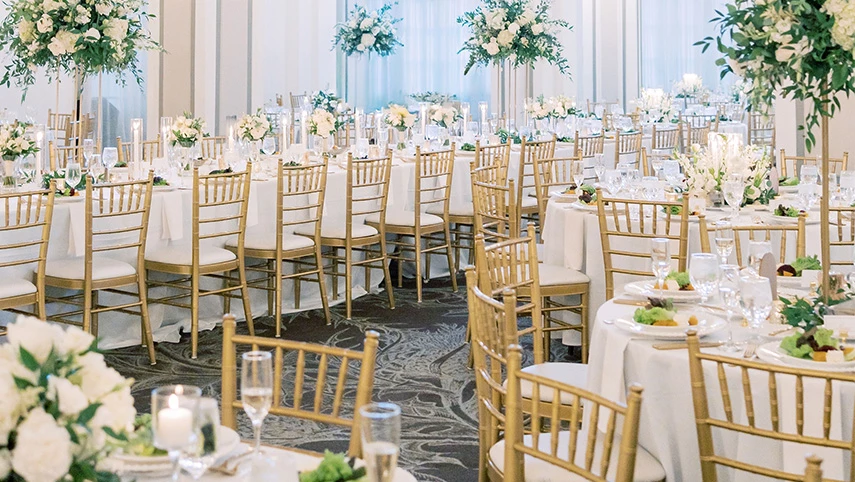 A large, elegantly set dining room with round tables, gold chairs, and floral centerpieces.