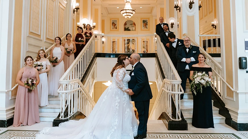 A wedding party stands on a grand staircase in a luxurious lobby