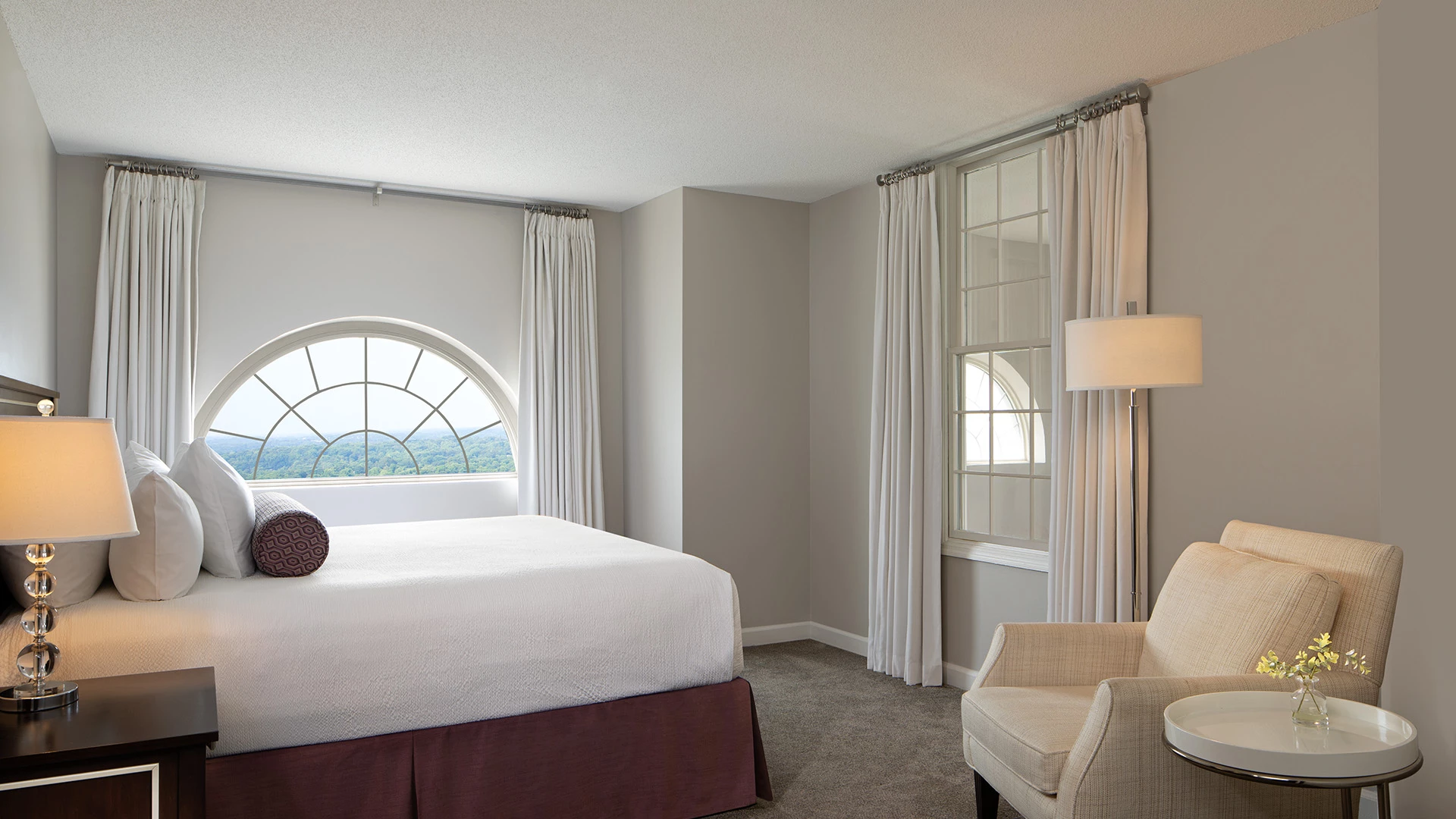 A hotel room with a bed, chair, and lamp in front of a window.
