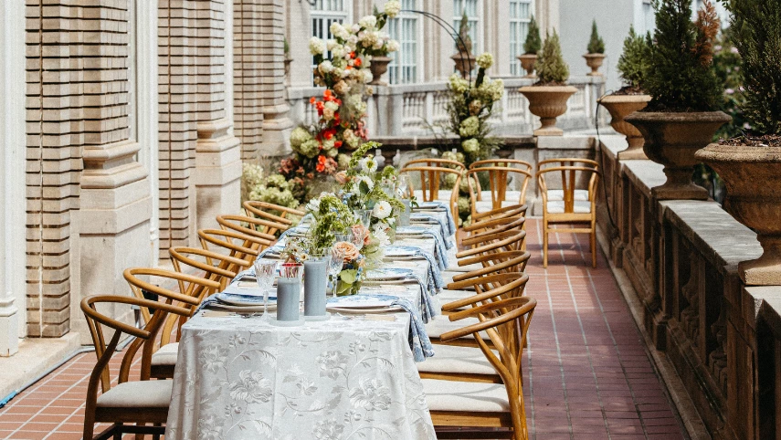 A long table set for a meal on a balcony with a white tablecloth, flowers, and wooden chairs.
