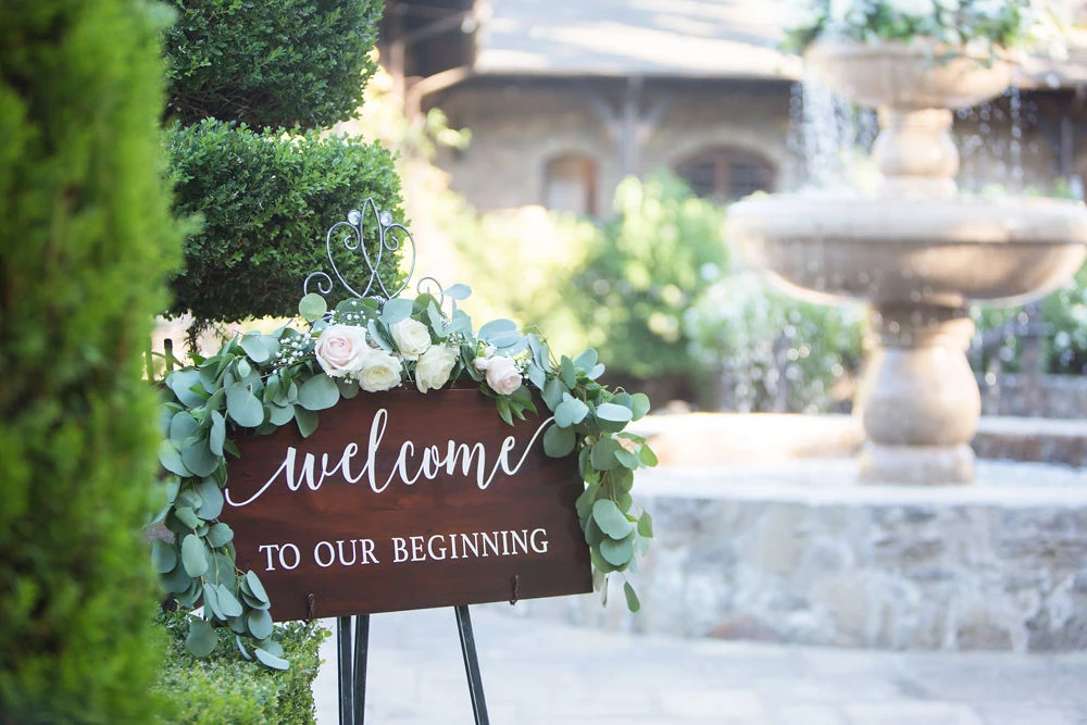 a sign with flowers and a fountain in the background
