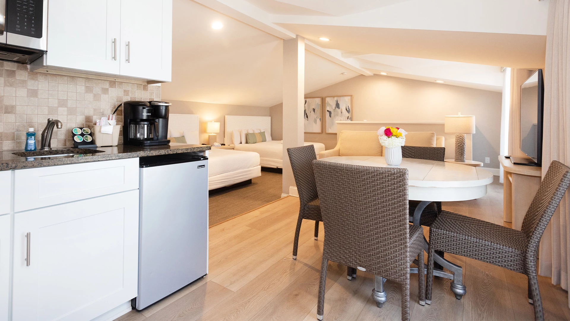 A wide view of a suite with a kitchenette, dining table, sofa, beds, and TV