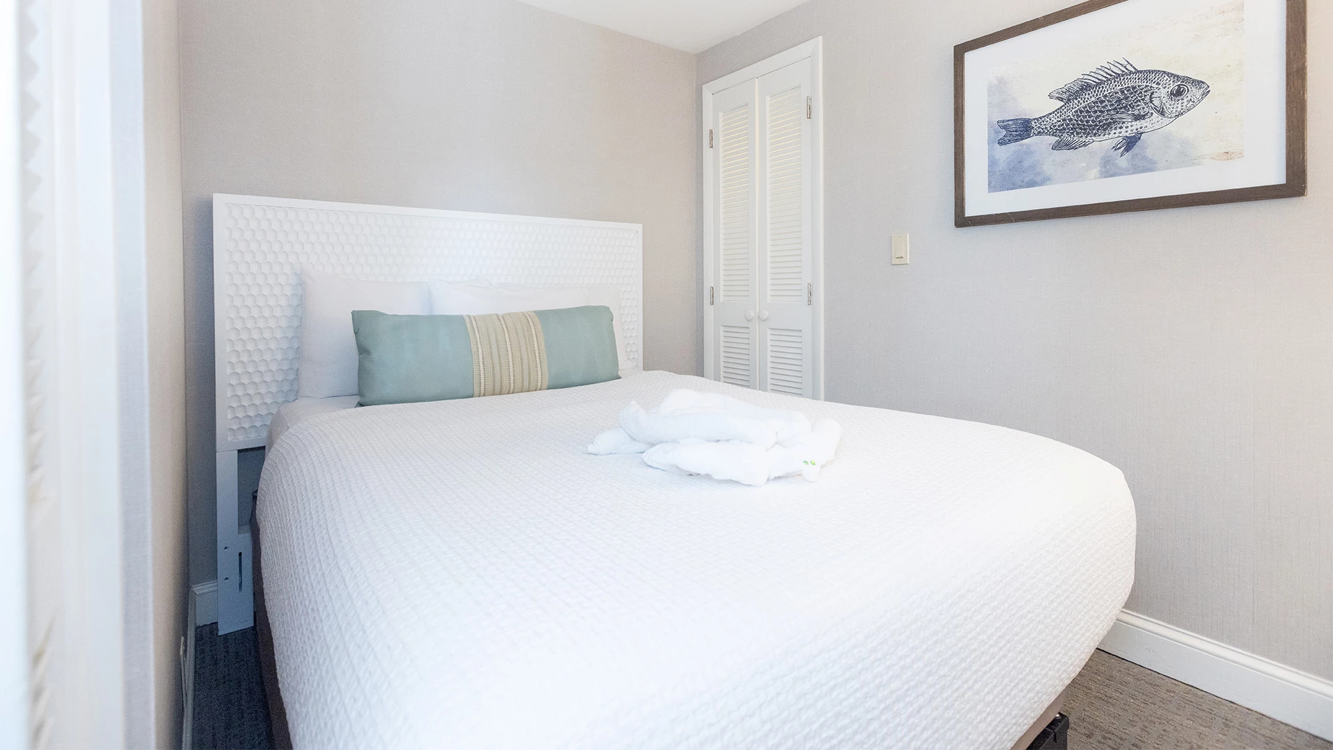 A neatly made bed with white linens and two pillows, one with a blue and beige design, in a room with light gray walls and a framed picture of a fish.