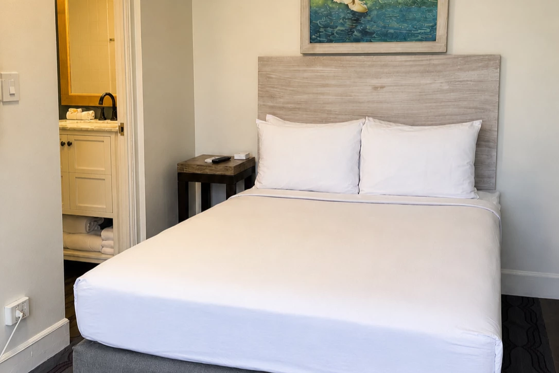 A hotel room with a bed, nightstand, and painting, with a bathroom doorway in the background.